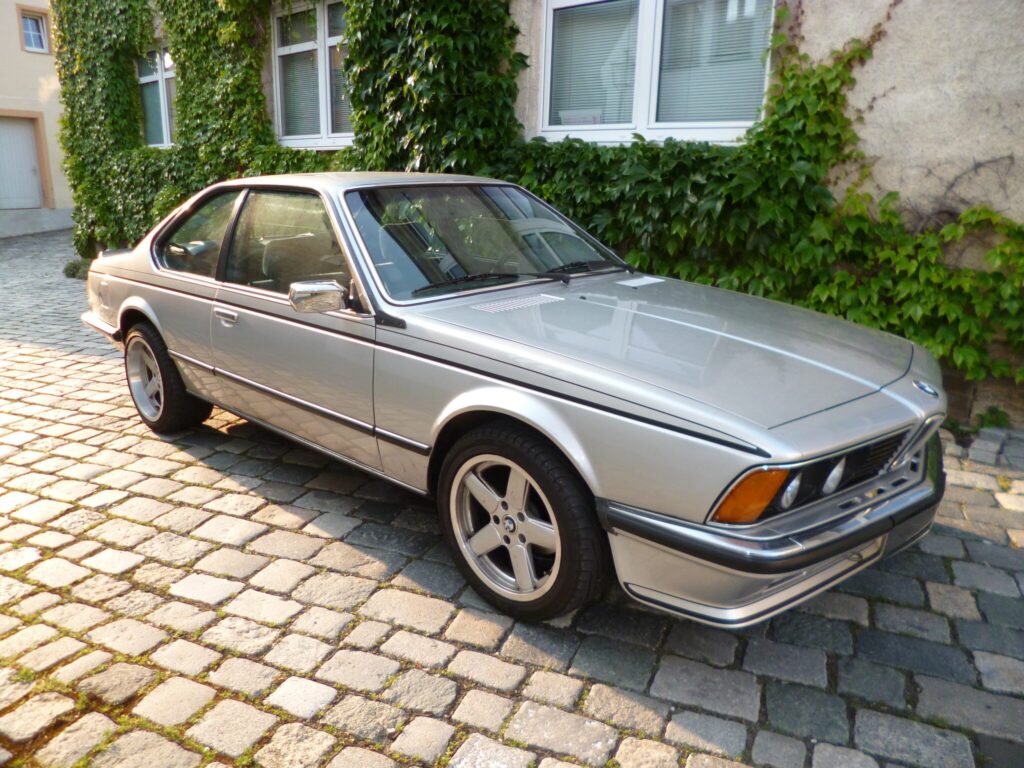Silberner BMW E24 auf Kopfsteinpflaster vor einer efeubewachsenen Fassade
